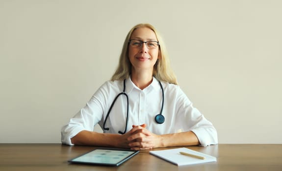 Caucasian middle-aged woman doctor with stethoscope, paper documents and digital tablet computer sitting at desk in clinic office
