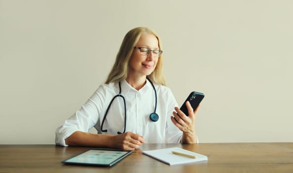 Caucasian middle-aged woman doctor using mobile phone with paper documents and digital tablet computer sitting at desk in clinic office