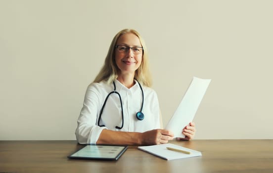 Caucasian middle-aged woman doctor with stethoscope, paper documents and digital tablet computer sitting at desk in clinic office