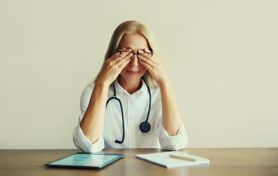 Tired caucasian middle-aged woman doctor working with digital tablet and paper documents while sitting at desk in medical clinic office