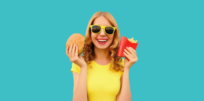 Portrait of happy cheerful young woman eating burger fast food and french fries, fried potatoes isolated on blue studio background