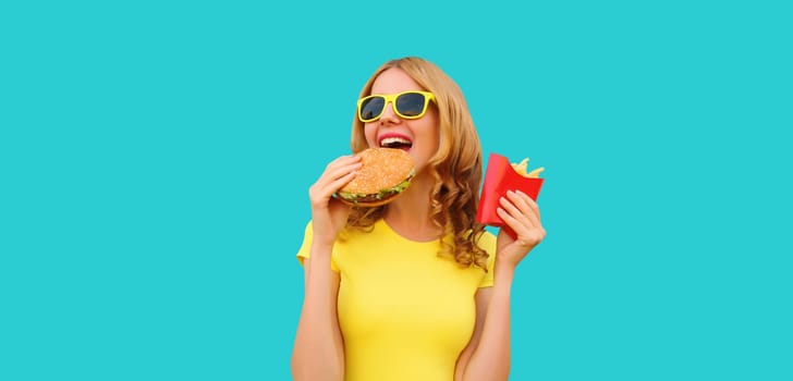 Portrait of happy cheerful young woman eating burger fast food and french fries, fried potatoes isolated on blue studio background