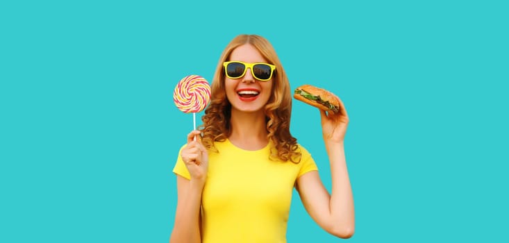 Portrait of happy cheerful young woman having fun with burger fast food and colorful sweet lollipop isolated on blue studio background