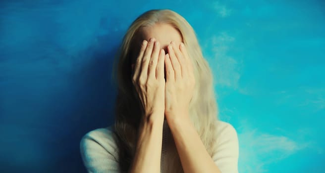 Upset sad crying middle-aged woman covering her eyes and face with her hands while experiencing mental or physical suffering sitting on couch in living room at home