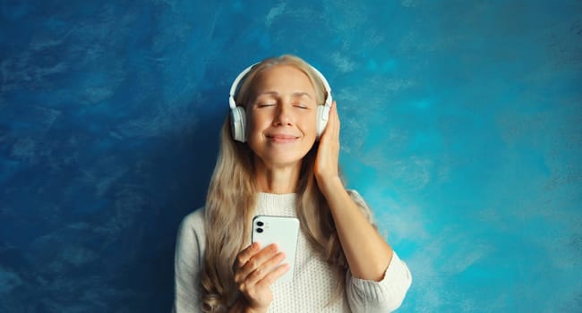 Portrait of happy smiling caucasian middle aged woman listening to music in headphones with smartphone on blue background