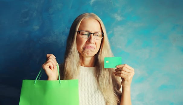Portrait of upset middle aged woman looking at plastic credit bank card with shopping bags on blue background