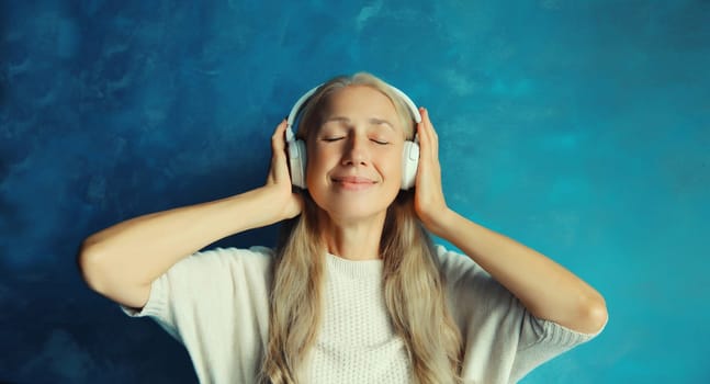 Portrait of happy smiling caucasian middle aged woman listening to music in wireless headphones on blue background