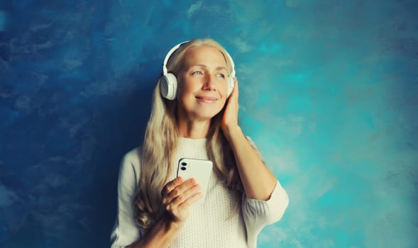 Portrait of happy smiling caucasian middle aged woman listening to music in headphones with smartphone on blue background