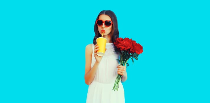 Portrait of beautiful woman with bouquet of red rose flowers drinking juice in heart shaped sunglasses on blue background
