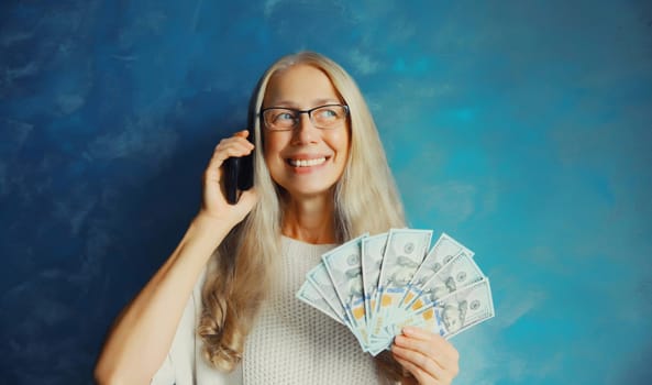 Portrait of happy caucasian smiling middle aged woman calling on mobile phone holding cash money in dollar bills in her hands