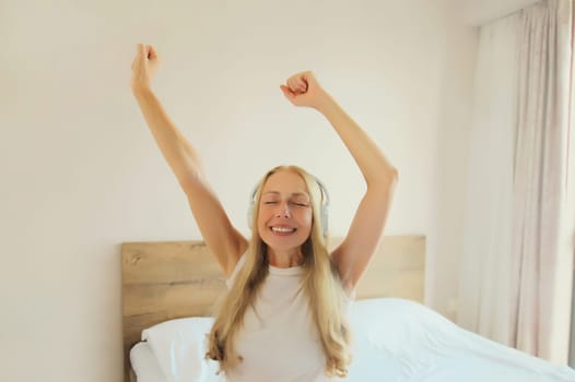Happy cheerful middle-aged woman listening to music with headphones having fun on the bed at home