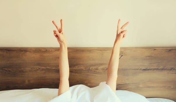Cheerful lazy woman waking up after sleeping lying in bed showing gesture stretching her hands up from under blanket in bedroom