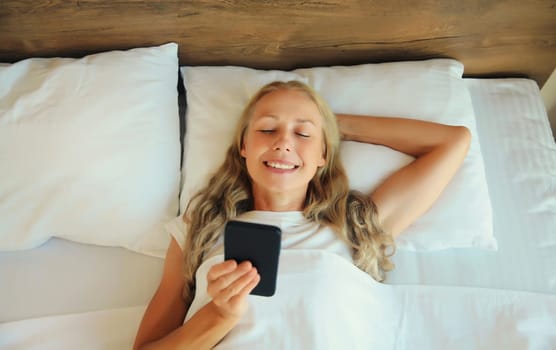 Healthy sleep, happy relaxed middle aged woman with mobile phone lying on the bed on white soft comfortable pillow in bedroom at home