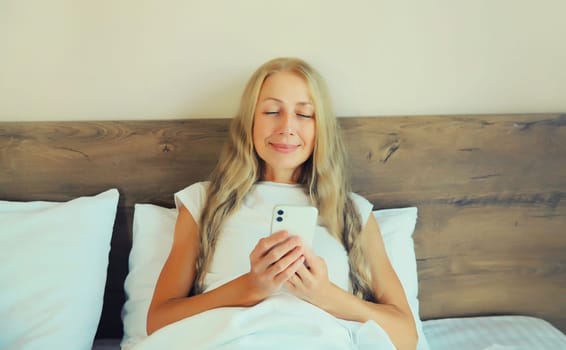 Healthy sleep, happy relaxed middle aged woman with mobile phone lying on the bed on white soft comfortable pillow in bedroom at home