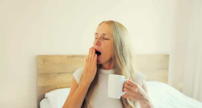 Lazy middle aged woman waking up after sleeping and yawning on bed holds cup of coffee in bedroom at home