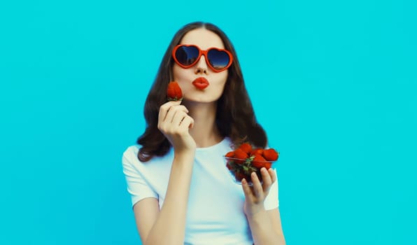 Portrait of beautiful caucasian young woman with handful of fresh strawberries blowing her lips wearing red heart shaped sunglasses on blue studio background