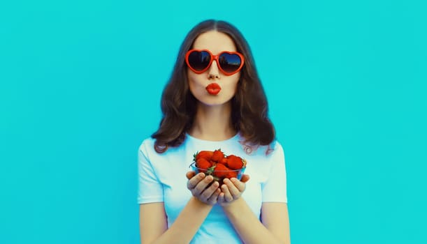 Portrait of beautiful caucasian young woman with handful of fresh strawberries blowing her lips wearing red heart shaped sunglasses on blue studio background
