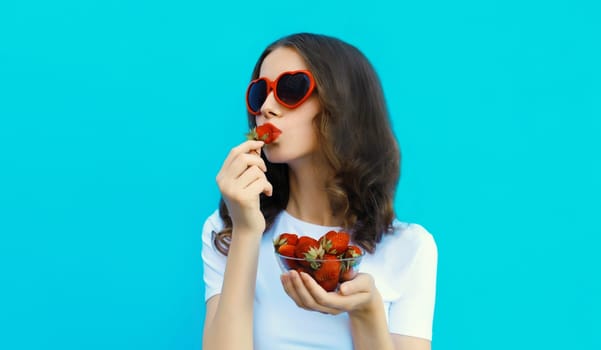 Portrait of beautiful caucasian young woman with handful of fresh strawberries blowing her lips wearing red heart shaped sunglasses on blue studio background