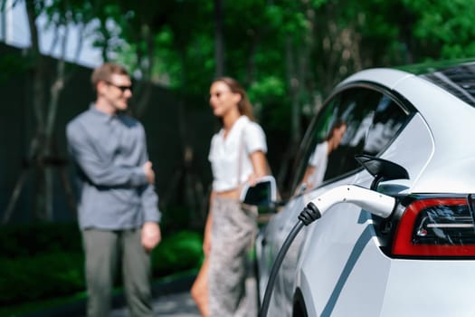 Young couple travel with EV electric car charging in green sustainable city outdoor garden in summer shows urban sustainability lifestyle by green clean rechargeable energy of electric vehicle innards