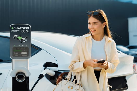 Young woman holding shopping bag and use smartphone to pay for electricity for recharging EV car battery from charging station at city mall parking lot. Modern woman go shopping by eco car. Expedient