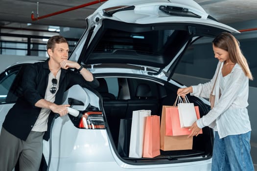 Young couple travel with EV electric car to shopping center parking lot charging in downtown city showing urban sustainability lifestyle by green clean rechargeable energy of electric vehicle innards