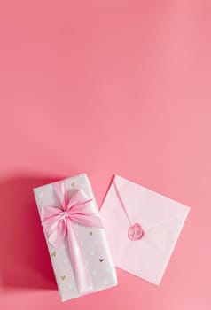 One beautiful gift box with a bow and a sealed envelope lie below on a pink background with copy space on top, flat lay close-up.