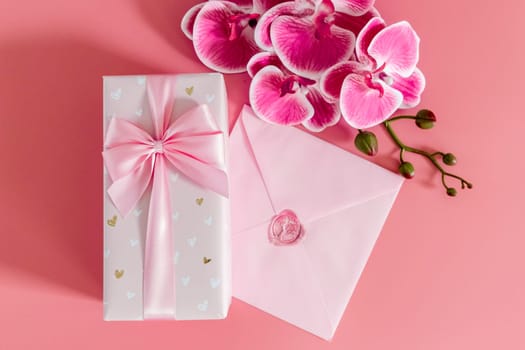 One beautiful gift box with a bow, a sealed envelope and orchid branches lie in the center on a pink background, flat lay close-up.