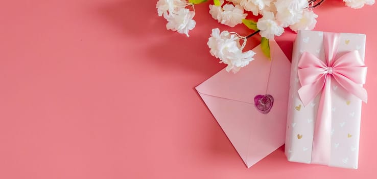 One beautiful gift box with a bow, a sealed envelope and branches of apple tree flowers lie on the right on a pink background with copy space on the left, flat lay close-up.