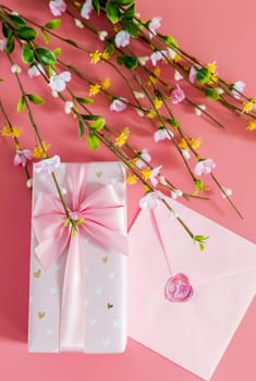One beautiful gift box with a bow, a sealed envelope and branches of spring flowers lie on a pink background, flat lay close-up with depth of field.