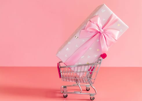 One large beautiful gift box with a bow lies in a mini shopping cart on the right on a pink background, close-up side view.