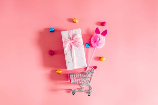 One large beautiful gift box with a bow, a fluffy bunny handle and chocolate Easter eggs in multi-colored wrappers fall out of a mini shopping cart on a pink background, flat lay close-up with copy space on the sides.