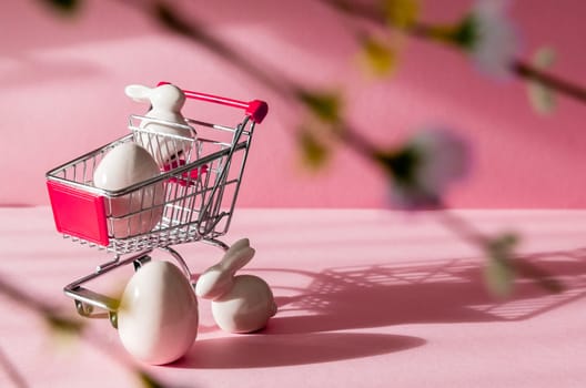 Porcelain figurines of Easter eggs and bunny with a mini shopping cart on the left on a pink background with shadows, blurred branches and spring flowers and copy space on the right, side view close-up.