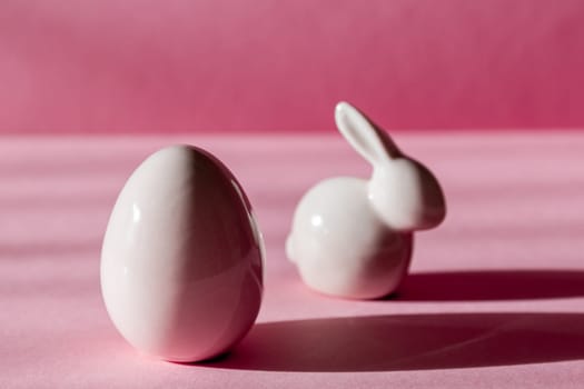 Porcelain figurines of Easter eggs and bunny stand on a pink background with shadows, close-up side view.