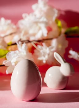 Porcelain figurines of Easter eggs and bunny stand on a pink background with a blurred branch of apple tree flowers, close-up side view.