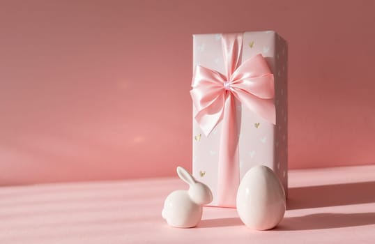 Porcelain figurines of Easter eggs and a big bunny gift box stand on the right on a pink background with shadows and copy space on the left, side view close-up.