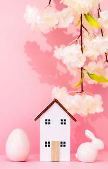 One wooden house with a porcelain Easter egg and a bunny stand on a pink background with a blurred branch of apple tree flowers, close-up side view with selective focus.