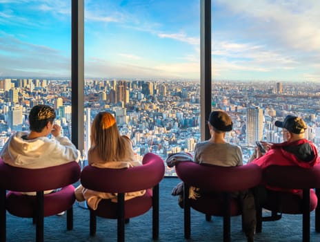 Back of caucasian tourists sitting on armchairs on the top floor of the observatory of the sightseeing Shibuya Sky admiring a Tokyo cityscape with its skyscrapers and buildings lit by the sunset light