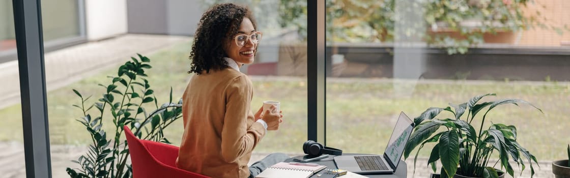 Smiling business woman in eyeglasses have a coffee break during working on laptop in cozy cafe