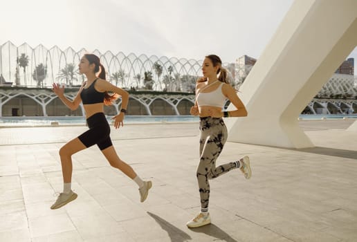 Two active women athlete running side by side along an outdoor track on modern buildings background
