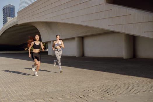 Two young women in sportswear are running on modern buildings background. Active lifestyle concept