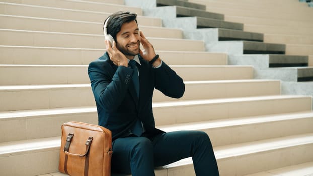 Professional business man picking and listening music by using headphone while sitting at stairs. Project manager dance while listening song while wearing headphone, suit. White background. Exultant.