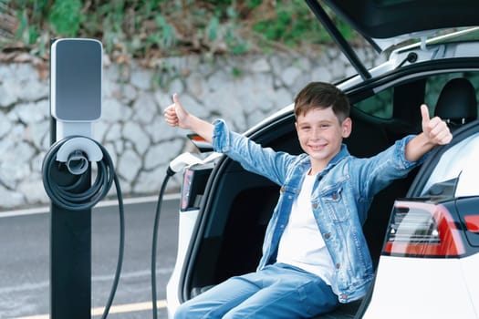 Little boy sitting on car trunk while recharging eco-friendly electric car from EV charging station. EV car road trip travel concept for alternative transportation powered sustainable energy.Perpetual