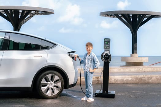 Little boy recharging eco-friendly electric car from EV charging station. EV car road trip travel by the seashore by alternative vehicle powered by clean renewable and sustainable energy. Perpetual