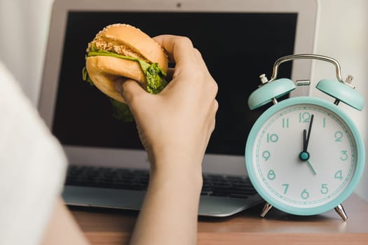 Person eating a hamburger while working, and there is an alarm clock displaying noon time on the work table for the concept of fast food eating at the workplace