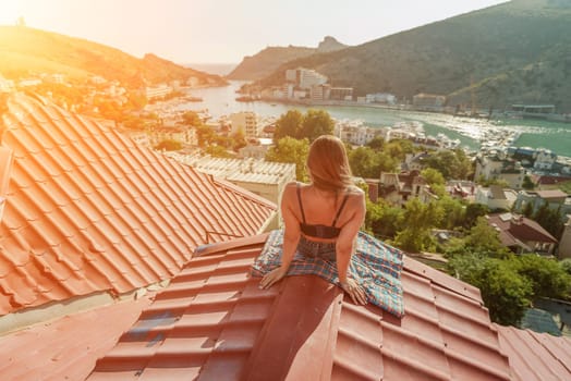 Woman sits on rooftop, enjoys town view and sea mountains. Peaceful rooftop relaxation. Below her, there is a town with several boats visible in the water. Rooftop vantage point