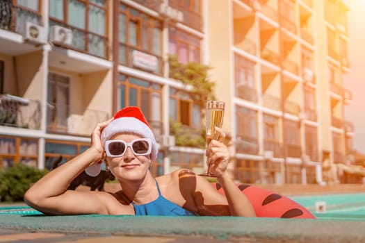 Woman pool Santa hat. A happy woman in a blue bikini, a red and white Santa hat and sunglasses poses near the pool with a glass of champagne standing nearby. Christmas holidays concept