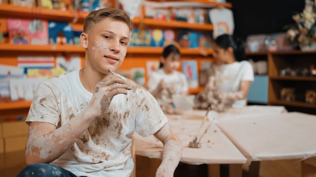 Happy student looking while put paintbrush behind ear in art lesson. Diverse student having pottery class together. Highschool boy smile while wearing white shirt with mud stained cloth. Edification.