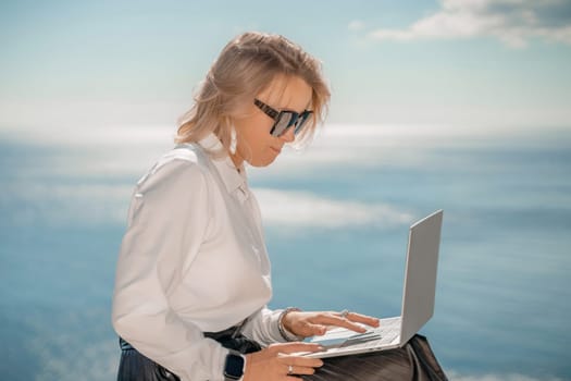 Business woman on nature in white shirt and black skirt. She works with an iPad in the open air with a beautiful view of the sea. The concept of remote work