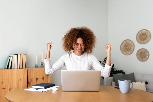 Happy young multiracial Hispanic female celebrating success with arms raised up in front of laptop at home office. Achievement concept.