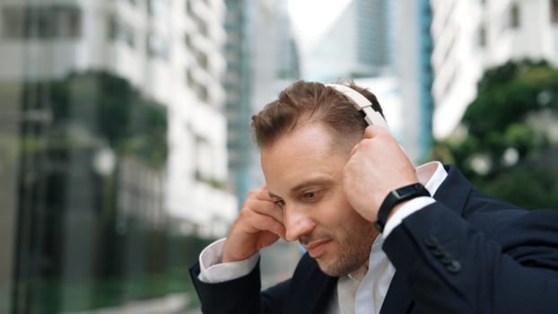 Businessman put on headphone and listen relaxing music while sitting at green city with blurred background . Skilled manager project manager enjoy to listen while dance to song in lively mood. Urbane.